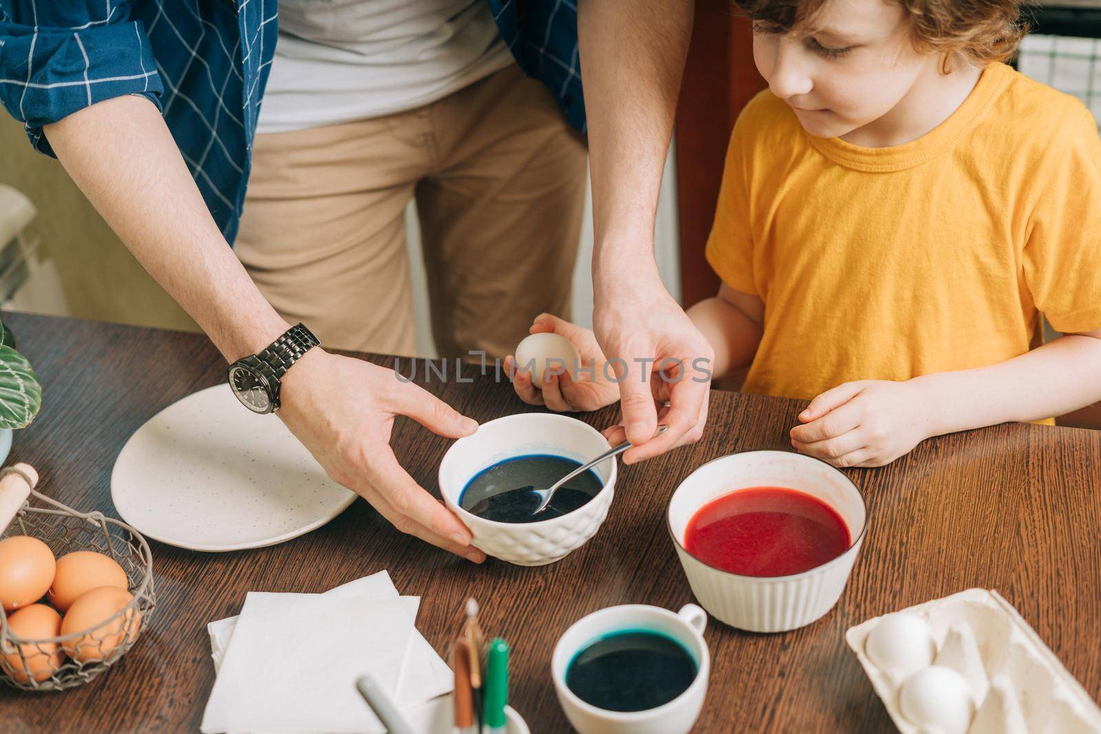 Easter day. Male Father and son painting eggs on wooden background. Family sitting in a kitchen. Preparing for Easter, creative homemade decoration. Child kid boy having fun and painting easter eggs