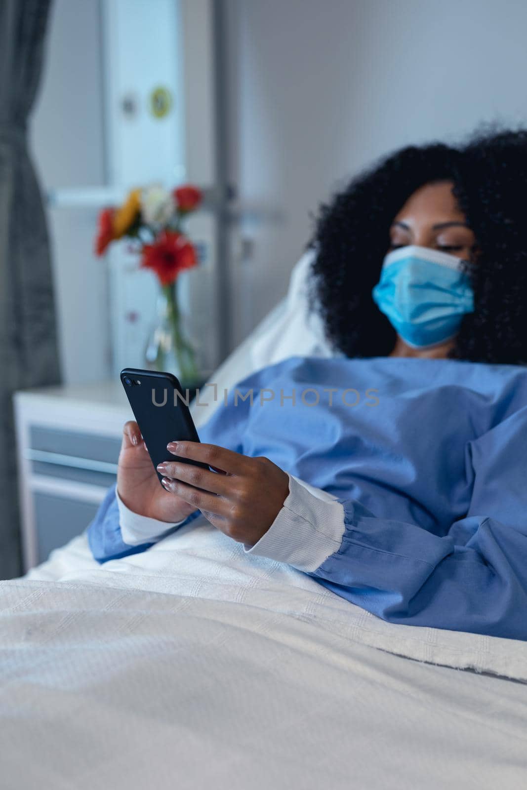 African american female patient lying in hospital bed wearing face mask using smartphone. medicine and health services during coronavirus covid 19 pandemic.