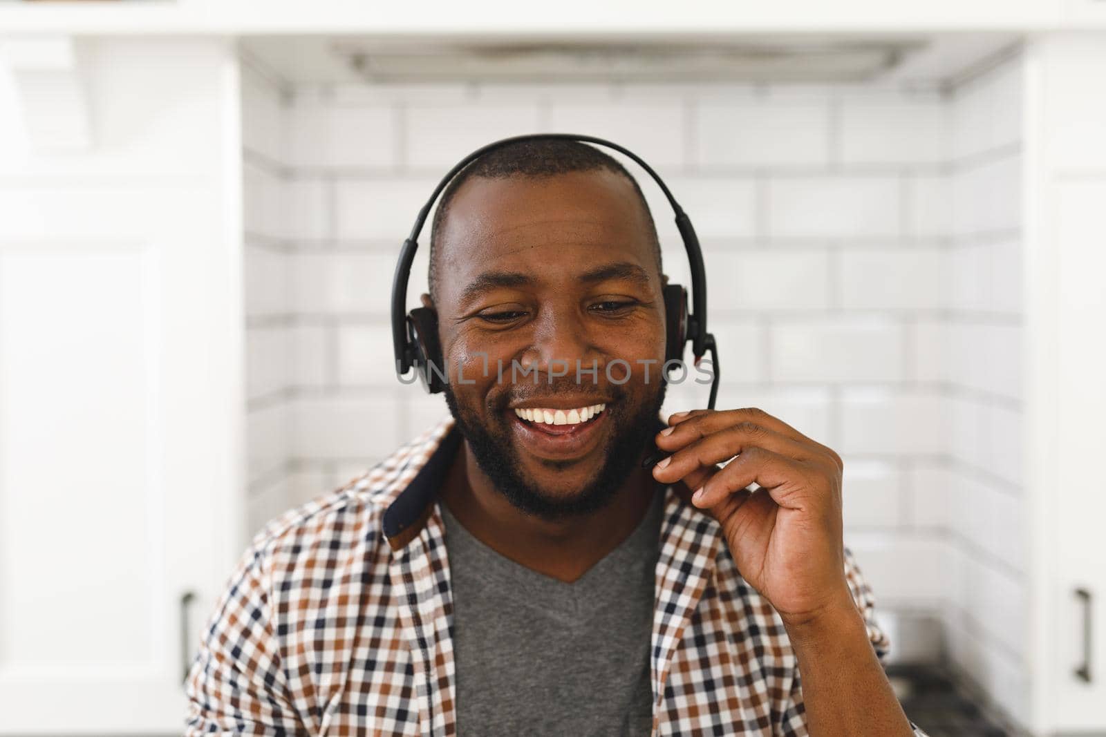 Smiling african american man sitting at countertop in kitchen making video call using headset. remote working from home with technology.