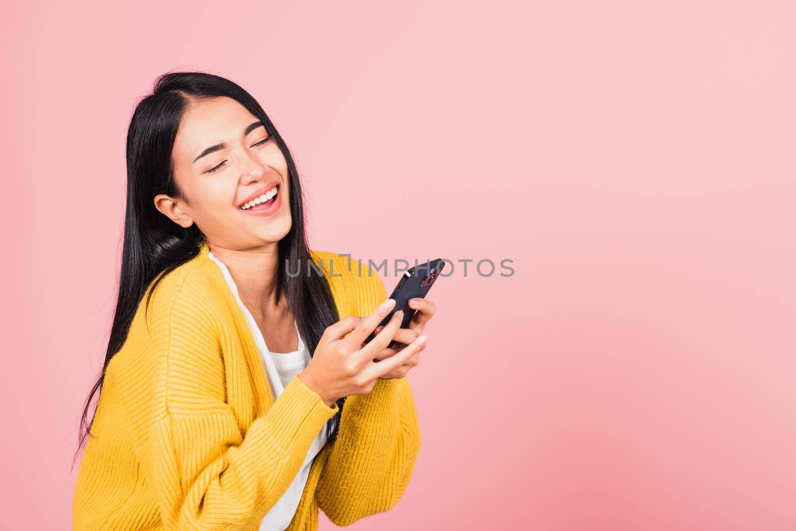 Happy Asian portrait beautiful cute young woman excited laughing holding mobile phone, studio shot isolated on pink background, female using funny smartphone making winner gesture