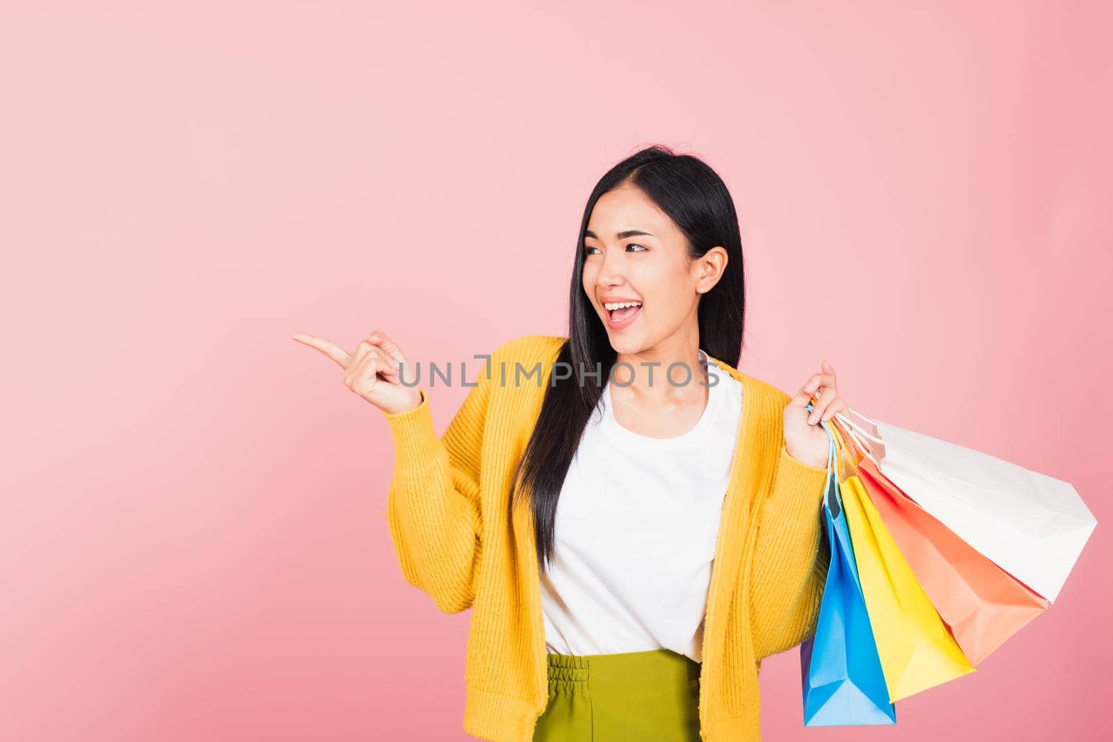 Portrait of Asian happy beautiful young woman shopper smiling standing excited holding online shopping bags multicolor in summer pointing finger to copy space, studio shot isolated on pink background