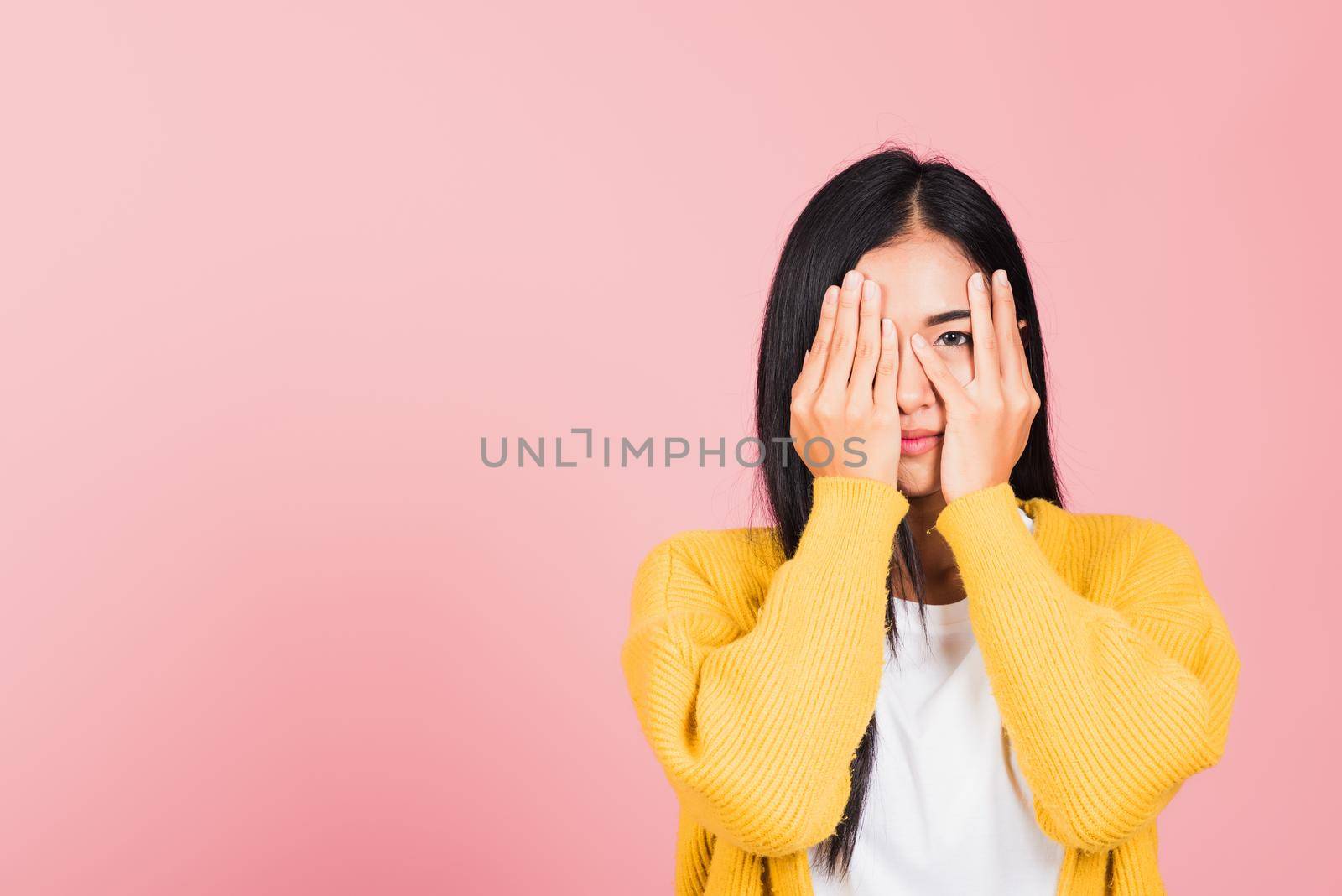 Asian portrait beautiful young woman in depressed bad mood covering face with hands and peering out with one eye between her fingers studio shot isolated on pink background surprised and shocked