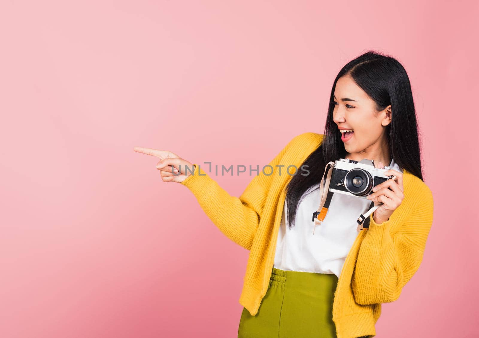 Attractive energetic happy Asian portrait beautiful cute young woman teen excited smiling holding vintage photo camera and pointing finger to side space, studio shot isolated on pink background