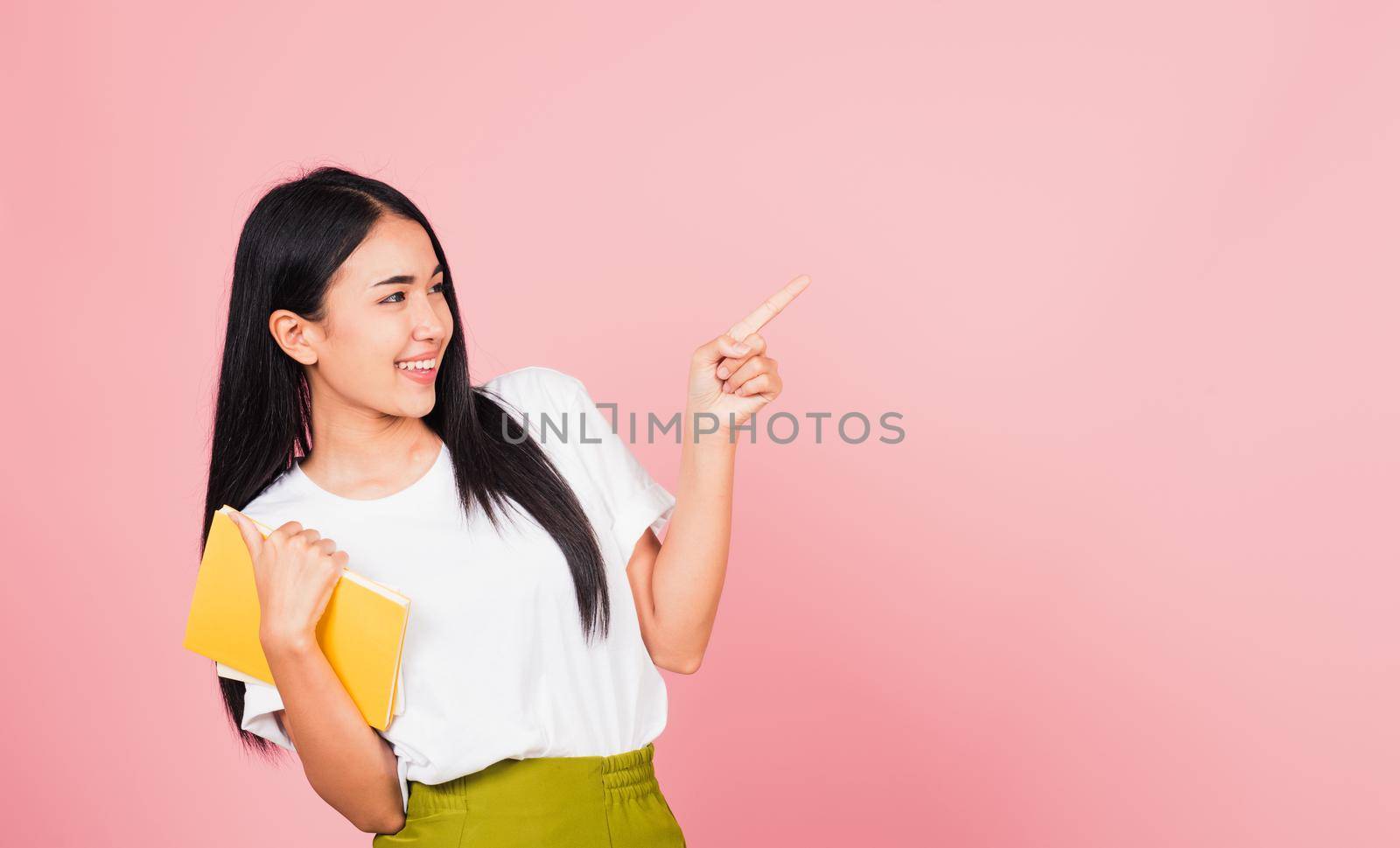 Portrait of happy Asian beautiful young woman confident smiling holding orange book open pointing finger to side copy space, studio shot isolated on pink background, education concept