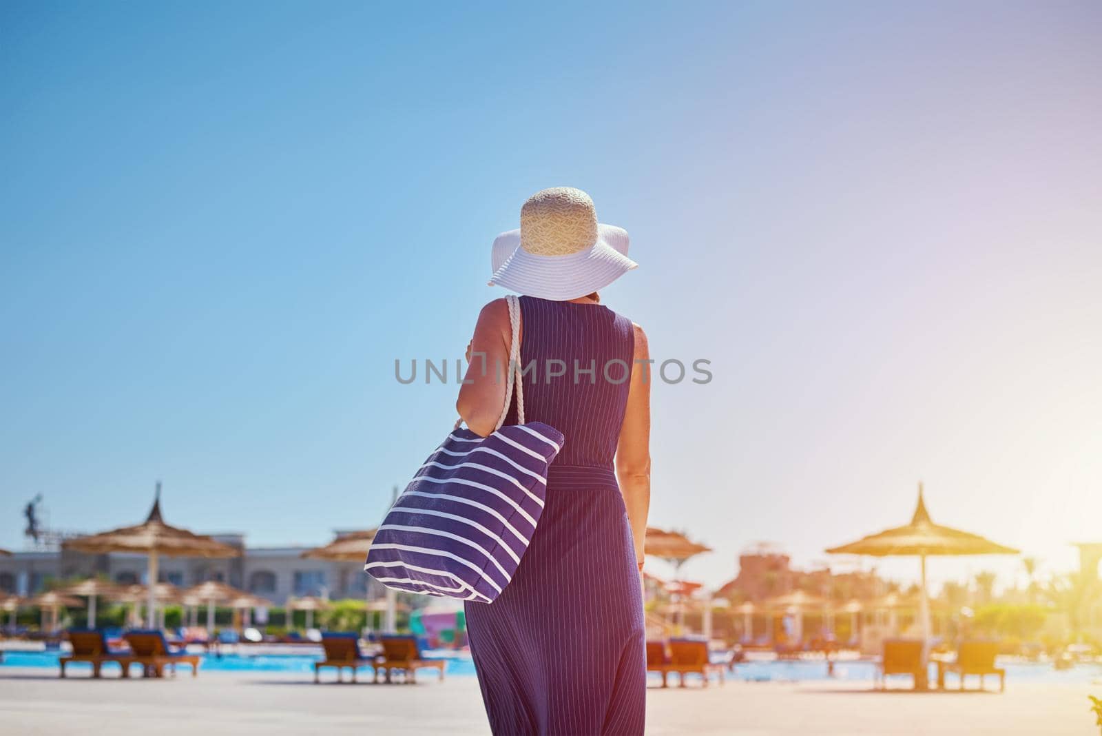 Woman in hat and blue dress go for a walk in the hotel resort near swimming pool with beach bag by Lazy_Bear