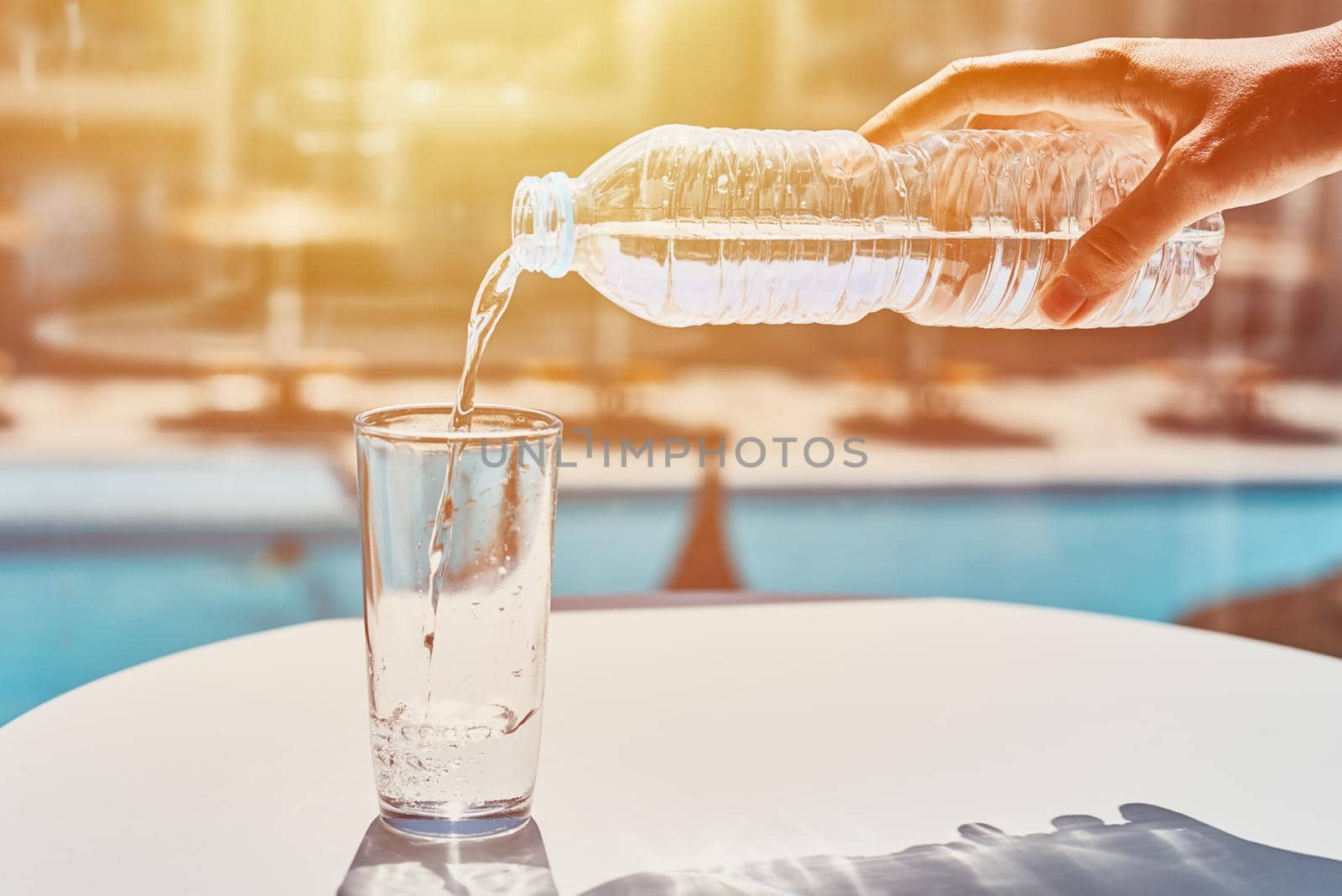 woman hand pouring water in glass from plastic bottle against swimming pool background