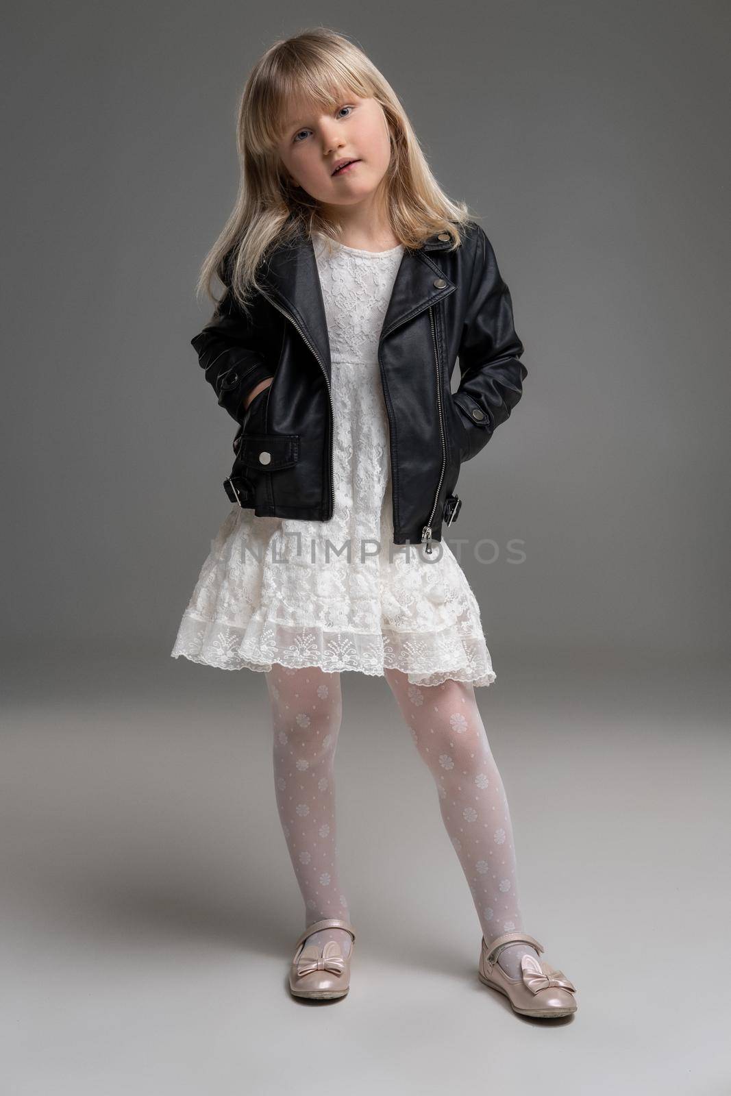 Little blonde girl in a white dress and black leather jacket is posing standing over a gray background. by nazarovsergey