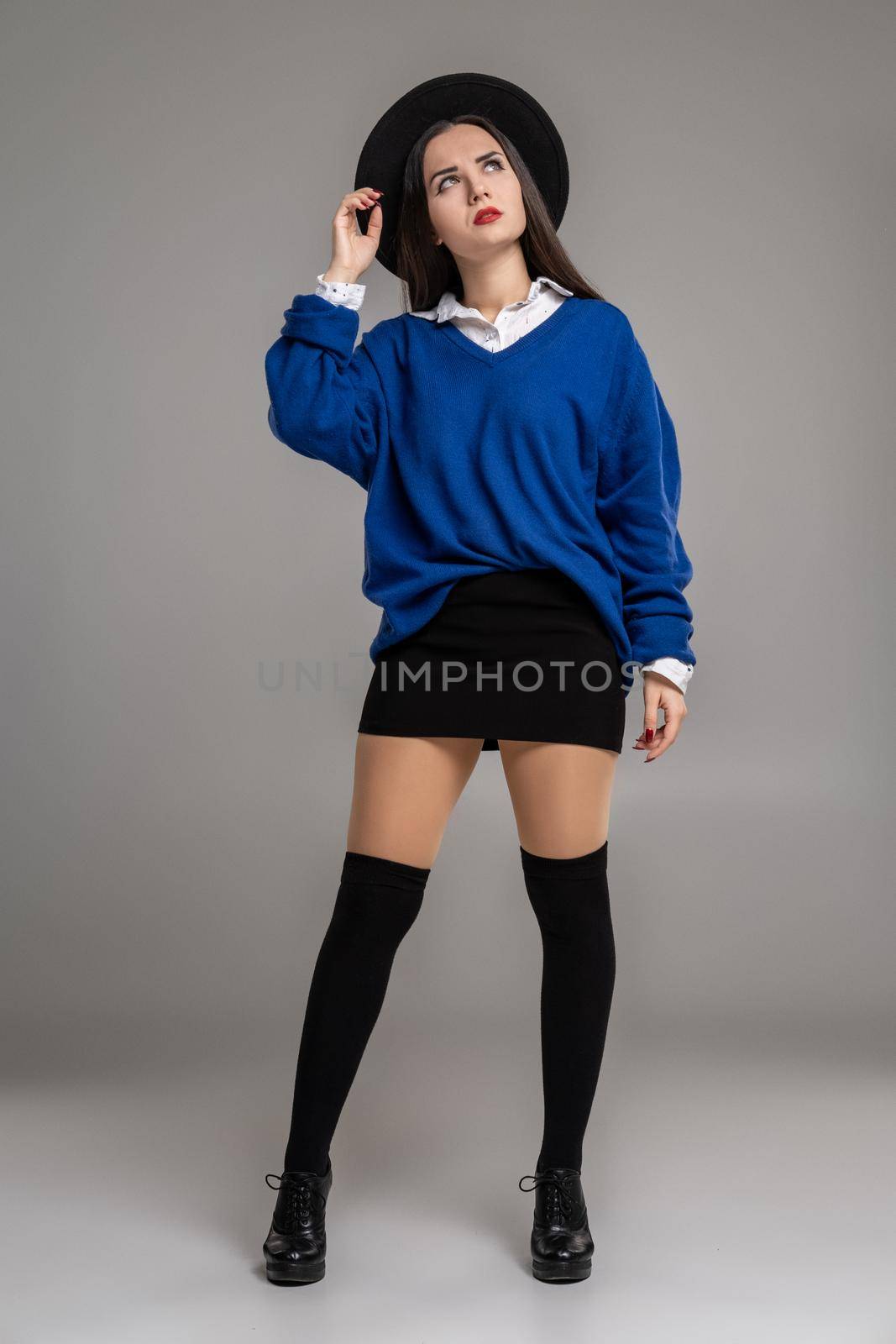 Full length portrait of an alluring girl holding her hat and looking up while posing at studio against a gray background. She is weared in a oversize blue blouse, black skirt, stockings and boots. Fashion shot. Sincere emotions concept.