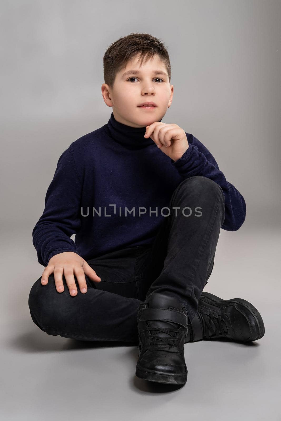 Full length portrait of a nice boy in a dark clothes and black boots sitting on the floor and looking at the camera while posing at studio as a fashion model. Photo of a schoolboy over a gray background. Copy space. Emotions concept.