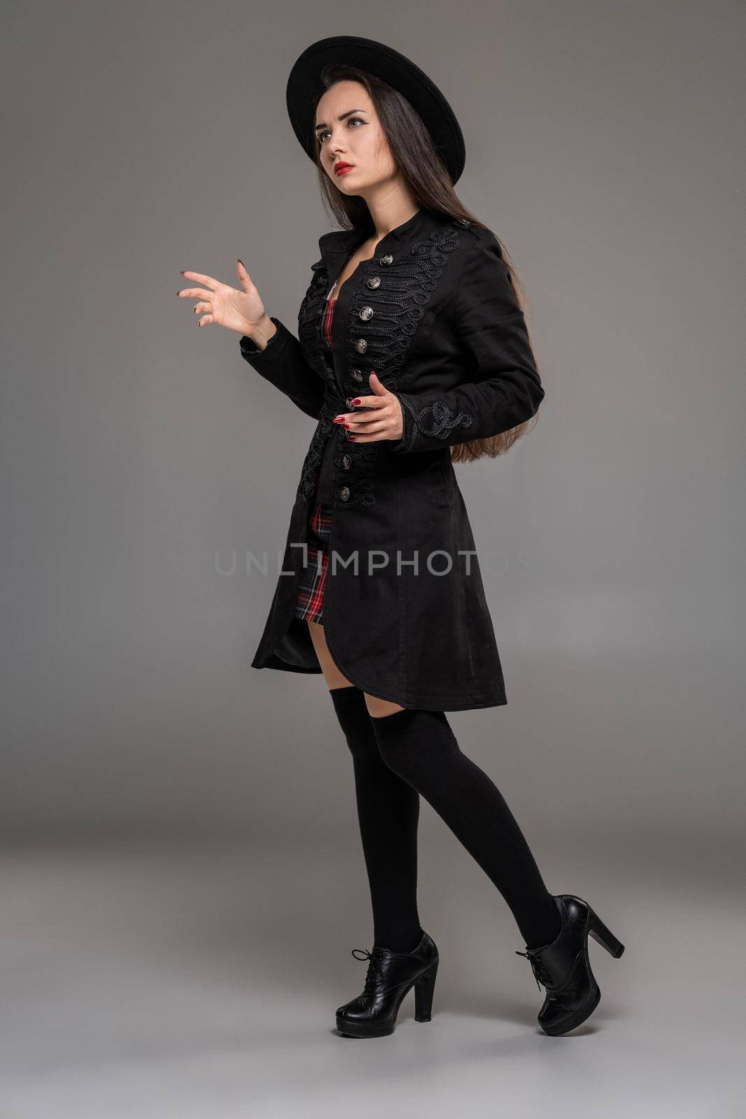 Full length portrait of a beautiful female posing sideways at studio against a gray background. She is weared in a checkered dress, black coat, hat, stockings and boots. Fashion shot. Sincere emotions concept.