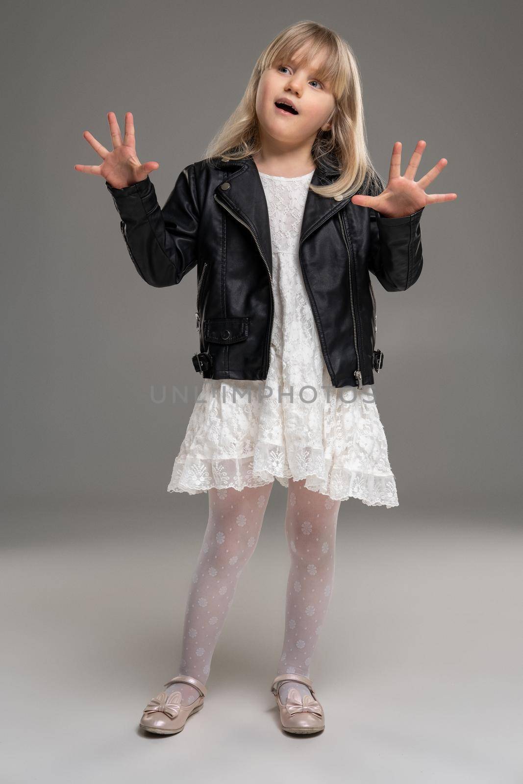 Full length photo of a pretty little blonde kid in a white dress and black leather jacket fooling around and posing standing over a gray background. Copy space. Sincere emotions concept.