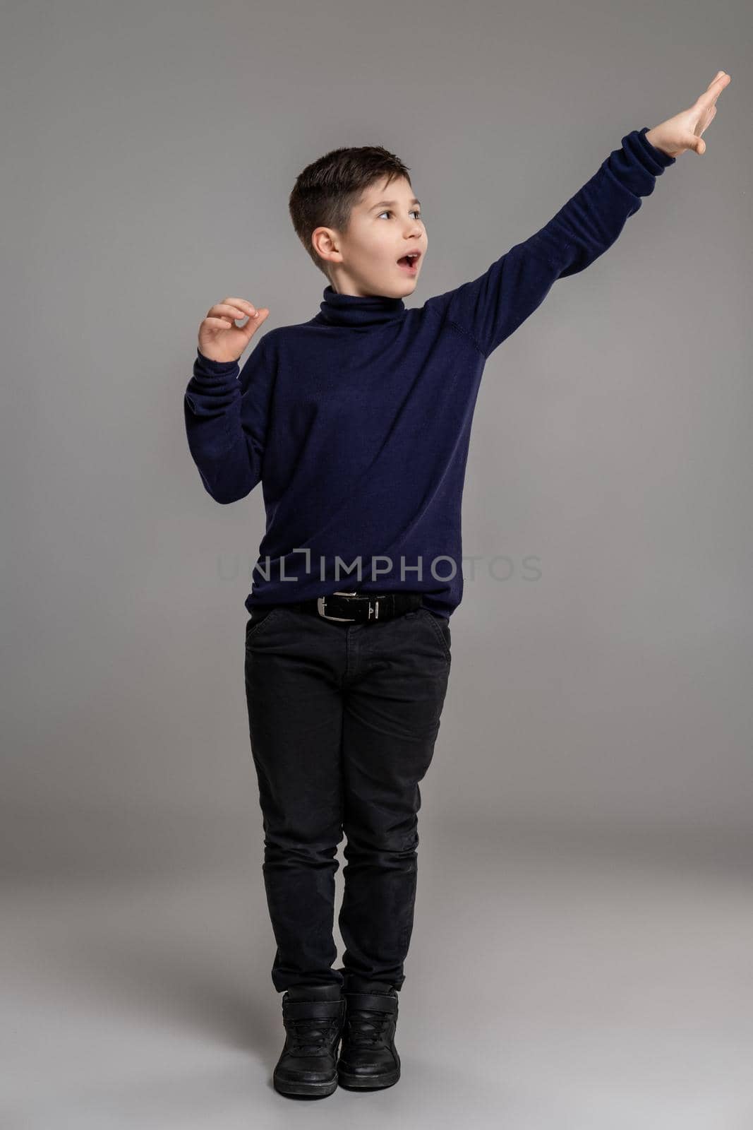 Full length portrait of a nice little man in a dark clothes and black boots gesticulating and looking away while posing at studio as a fashion model. Photo of a schoolboy over a gray background. Copy space. Emotions concept.