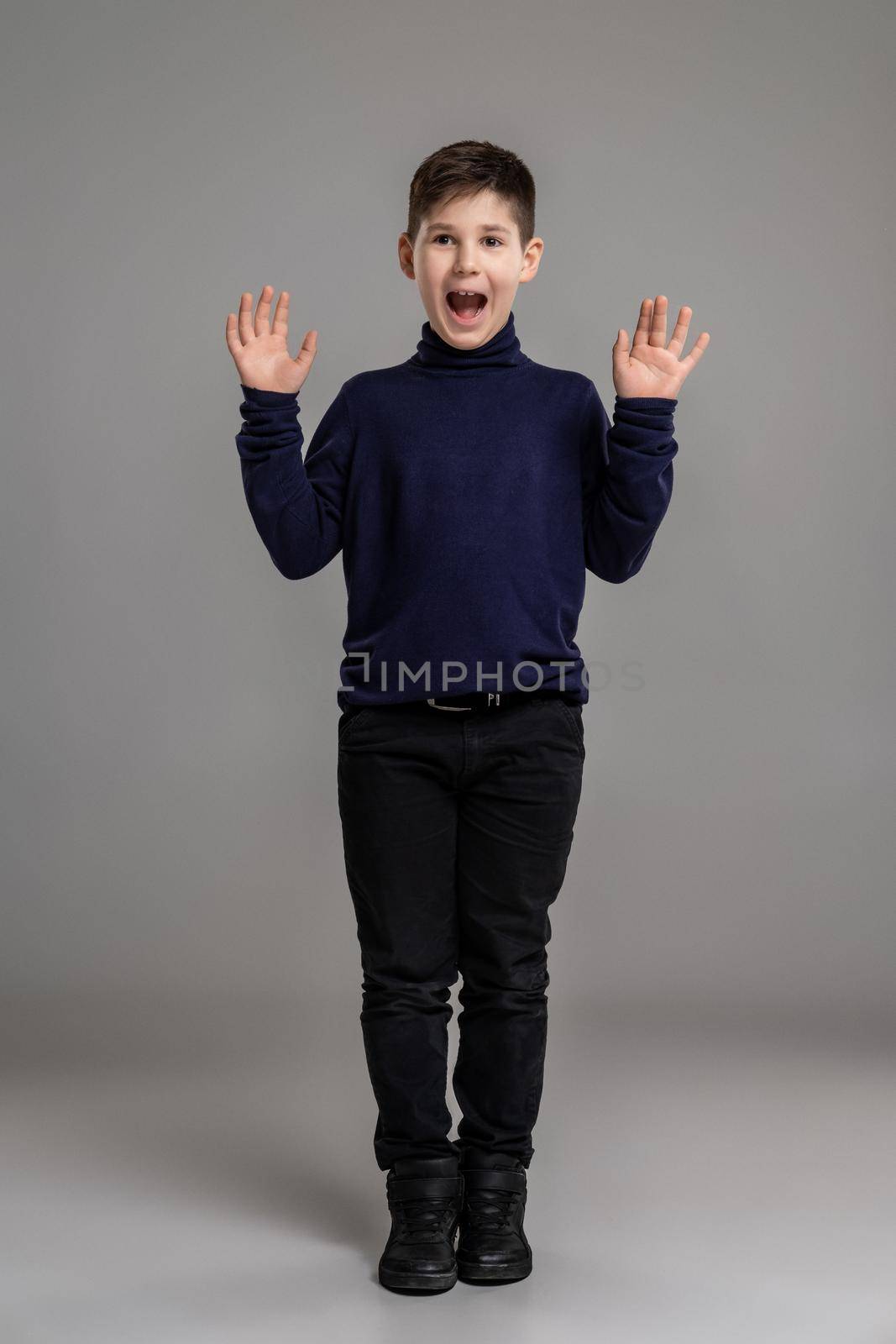 Full length portrait of a nice little male in a dark clothes and black boots screaming while posing at studio as a fashion model. Photo of a schoolboy over a gray background. Copy space. Emotions concept.