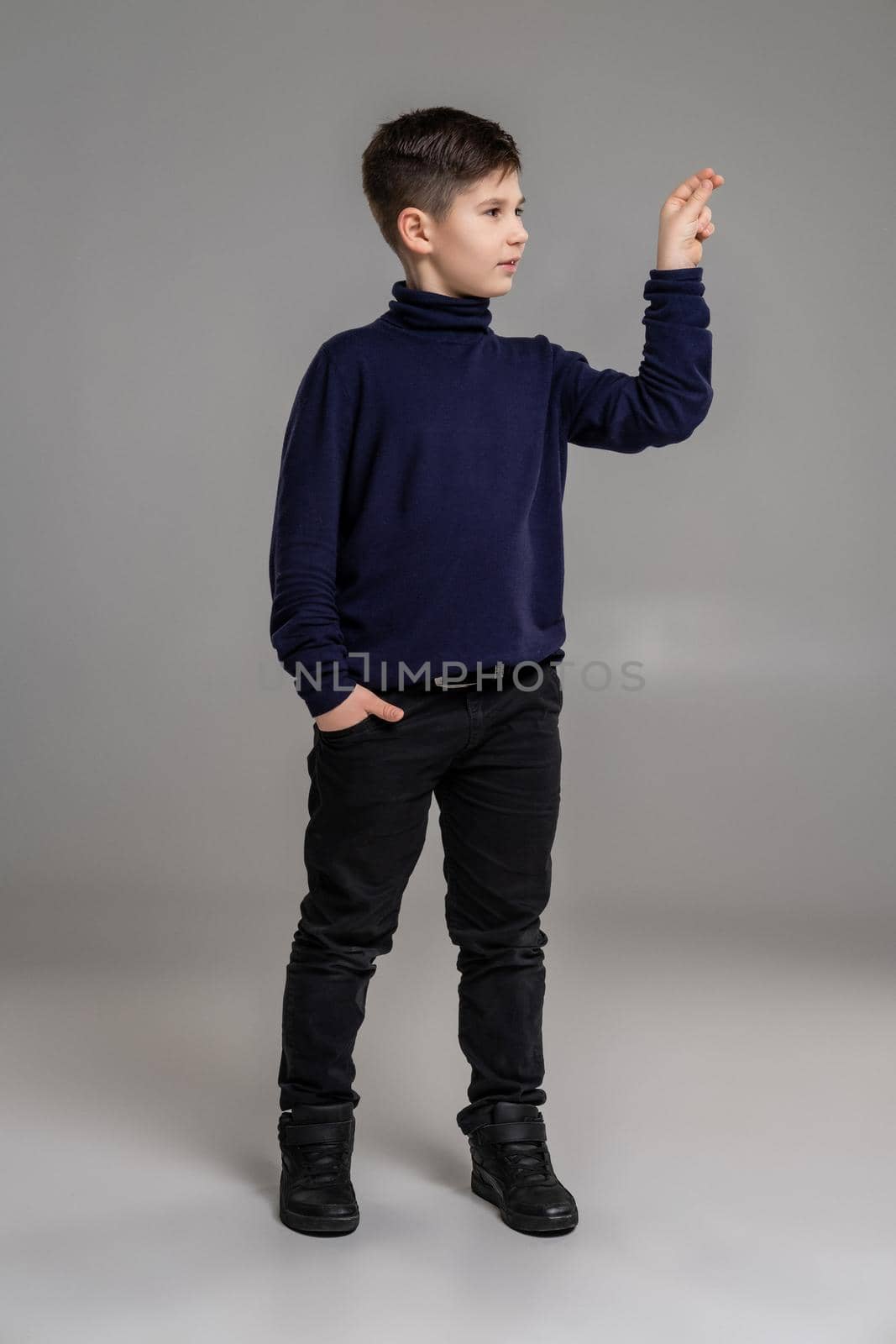 Full length portrait of a nice fellow in a dark clothes and black boots acting like holding something while posing at studio as a fashion model. Photo of a schoolboy over a gray background. Copy space. Emotions concept.