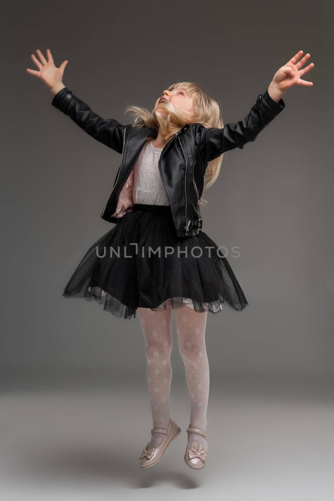 Little blonde girl in a white dress and black leather jacket is posing standing over a gray background. by nazarovsergey