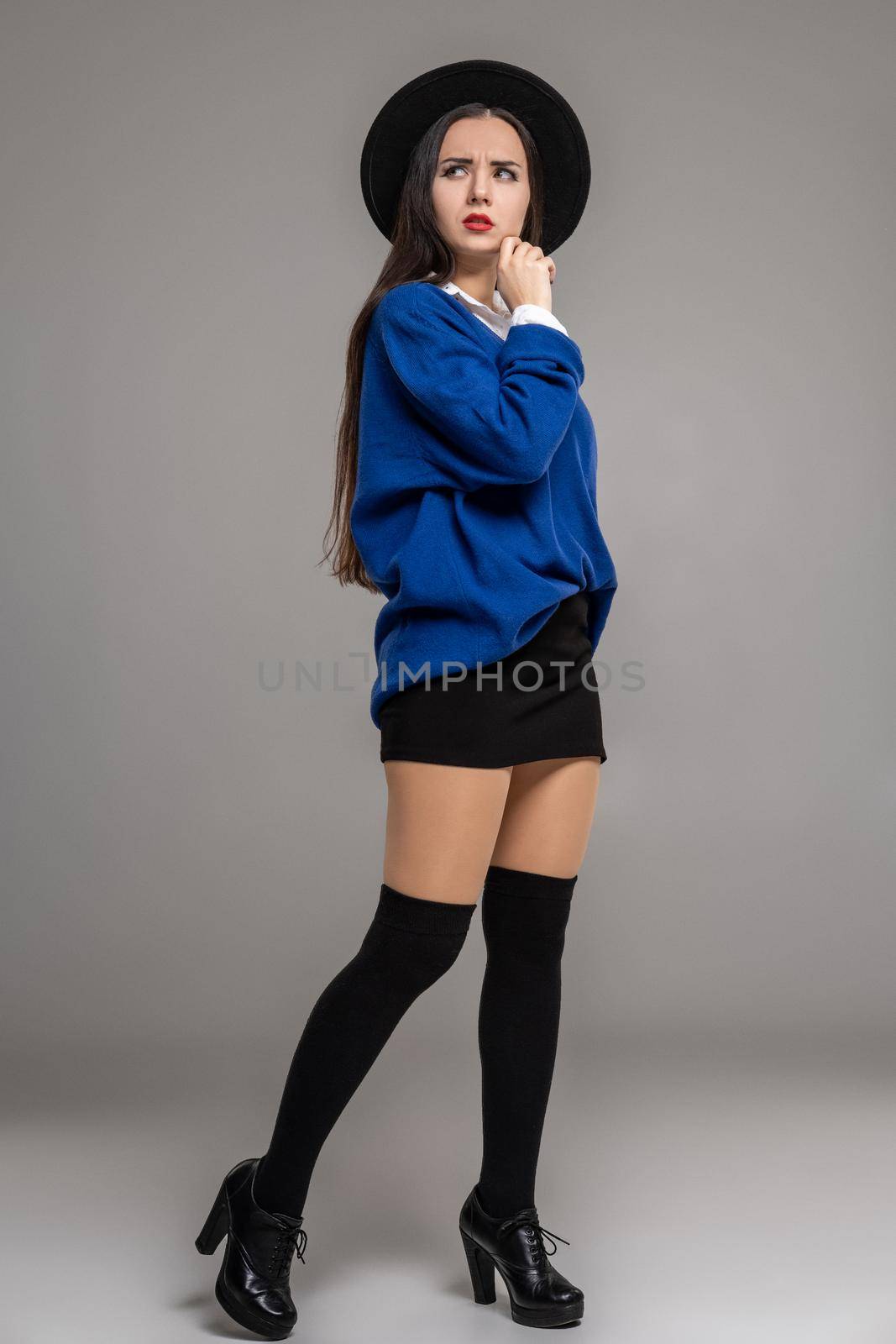 Full length portrait of an alluring woman posing at studio against a gray background. She is weared in a oversize blue blouse, black hat, skirt, stockings and boots. Fashion shot. Sincere emotions concept.