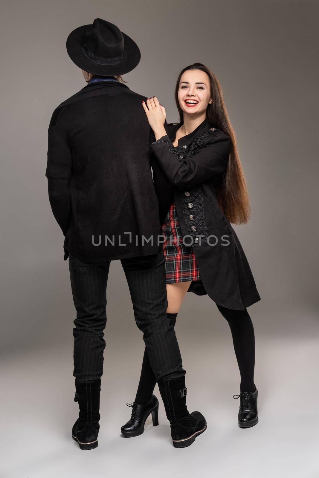 Young loving couple. Studio shot of a beautiful young maiden and man. They are in black outlet clothes and stylish hat posing over a gray studio background. Fashion shot. Sincere emotions concept. Copy space.