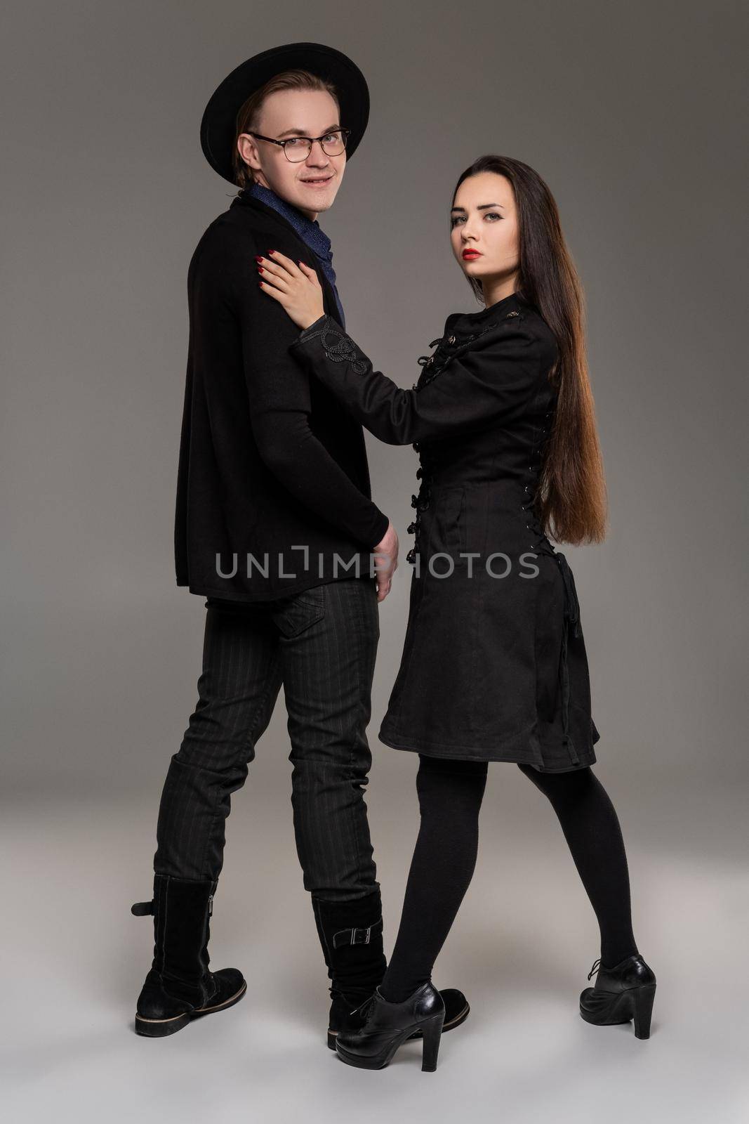 Young loving couple. Studio shot of a beautiful young lady and good-looking fellow. They are in black outlet clothes and stylish hat looking at the camera while posing over a gray studio background. Fashion shot. Sincere emotions concept. Copy space.