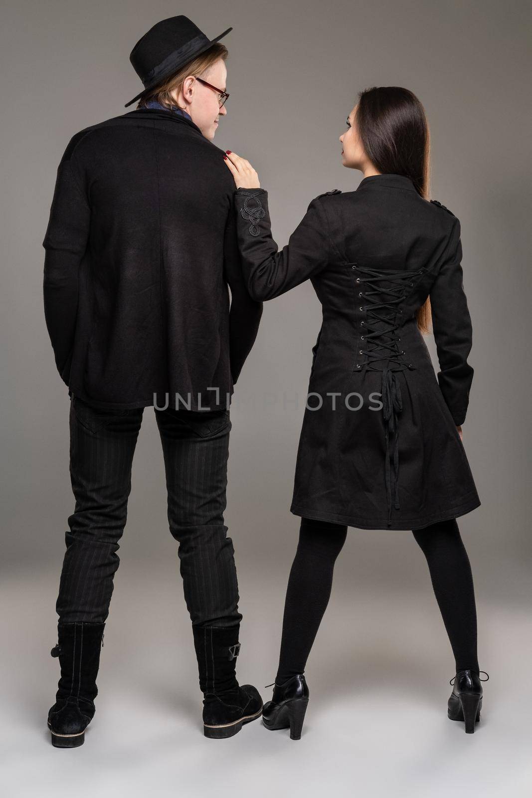 Young loving couple. Studio shot of a beautiful young female and handsome male. They are in black outlet clothes and stylish hat posing standing back to the camera over a gray studio background. Fashion shot. Sincere emotions concept. Copy space.