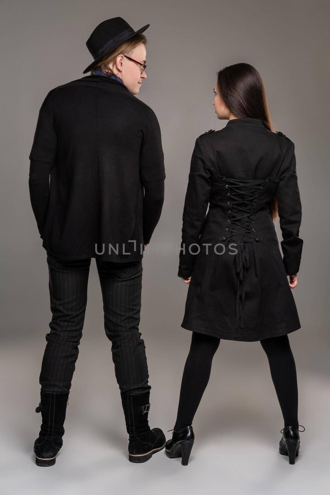 Young loving couple. Studio shot of a beautiful young girl and guy. They are in black outlet clothes and stylish hat posing standing back to the camera over a gray studio background. Fashion shot. Sincere emotions concept. Copy space.
