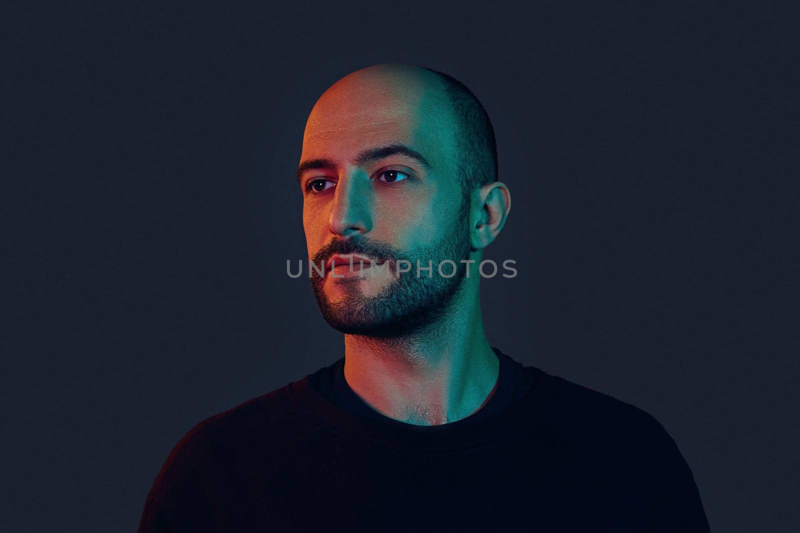 Epitome of masculinity. Portrait of a handsome dark-haired bearded young male in a black sweater looking away while posing against a colorful background. Modern and dramatic foto.