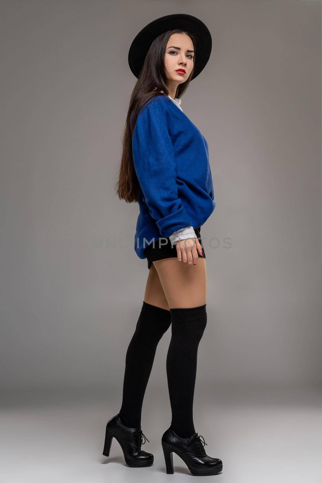 Full length portrait of an alluring lady posing sideways at studio against a gray background. She is weared in a oversize blue blouse, black hat, skirt, stockings and boots. Fashion shot. Sincere emotions concept.