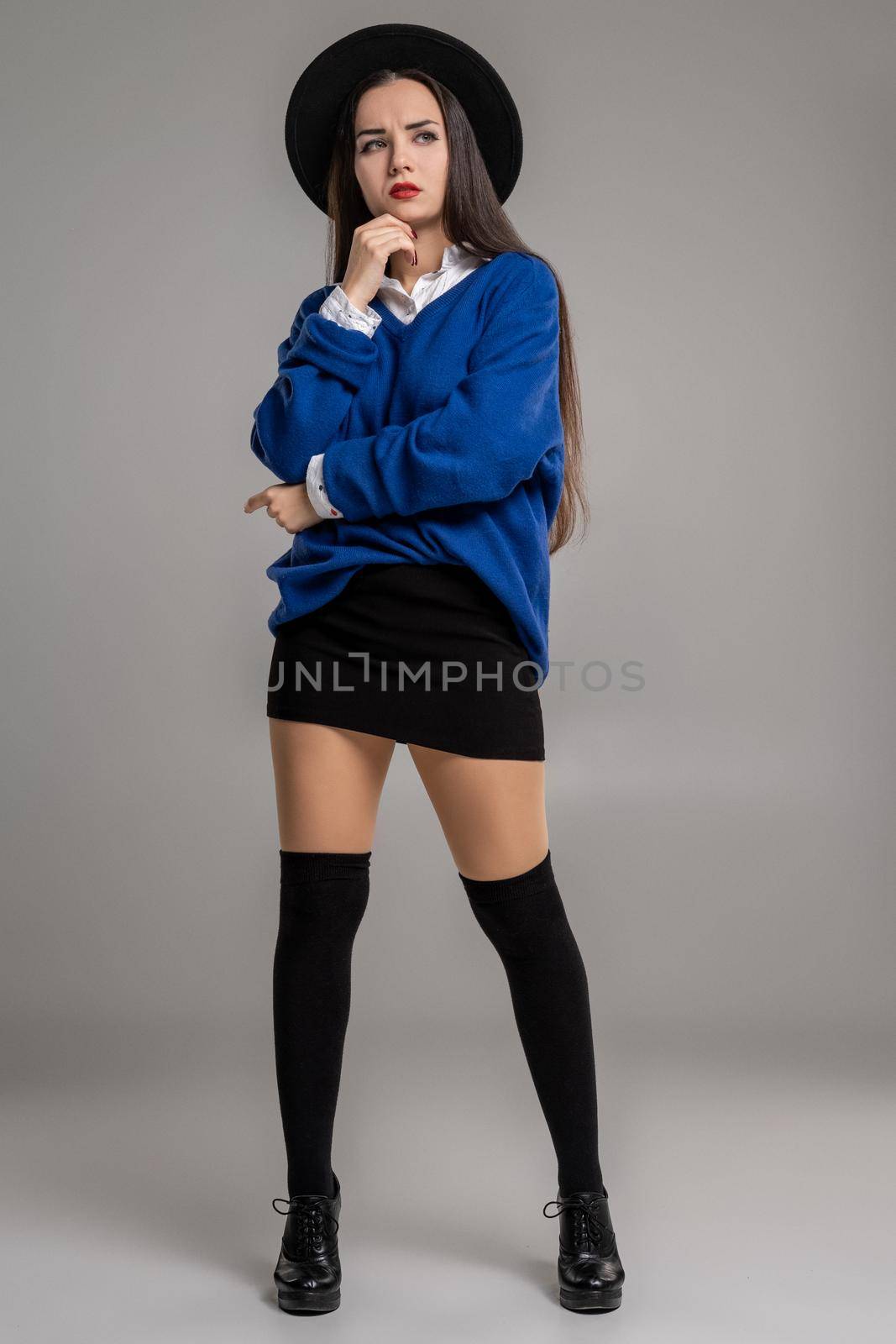 Full length portrait of an alluring female folded her hands and looking away while posing at studio against a gray background. She is weared in a oversize blue blouse, black hat, skirt, stockings and boots. Fashion shot. Sincere emotions concept.