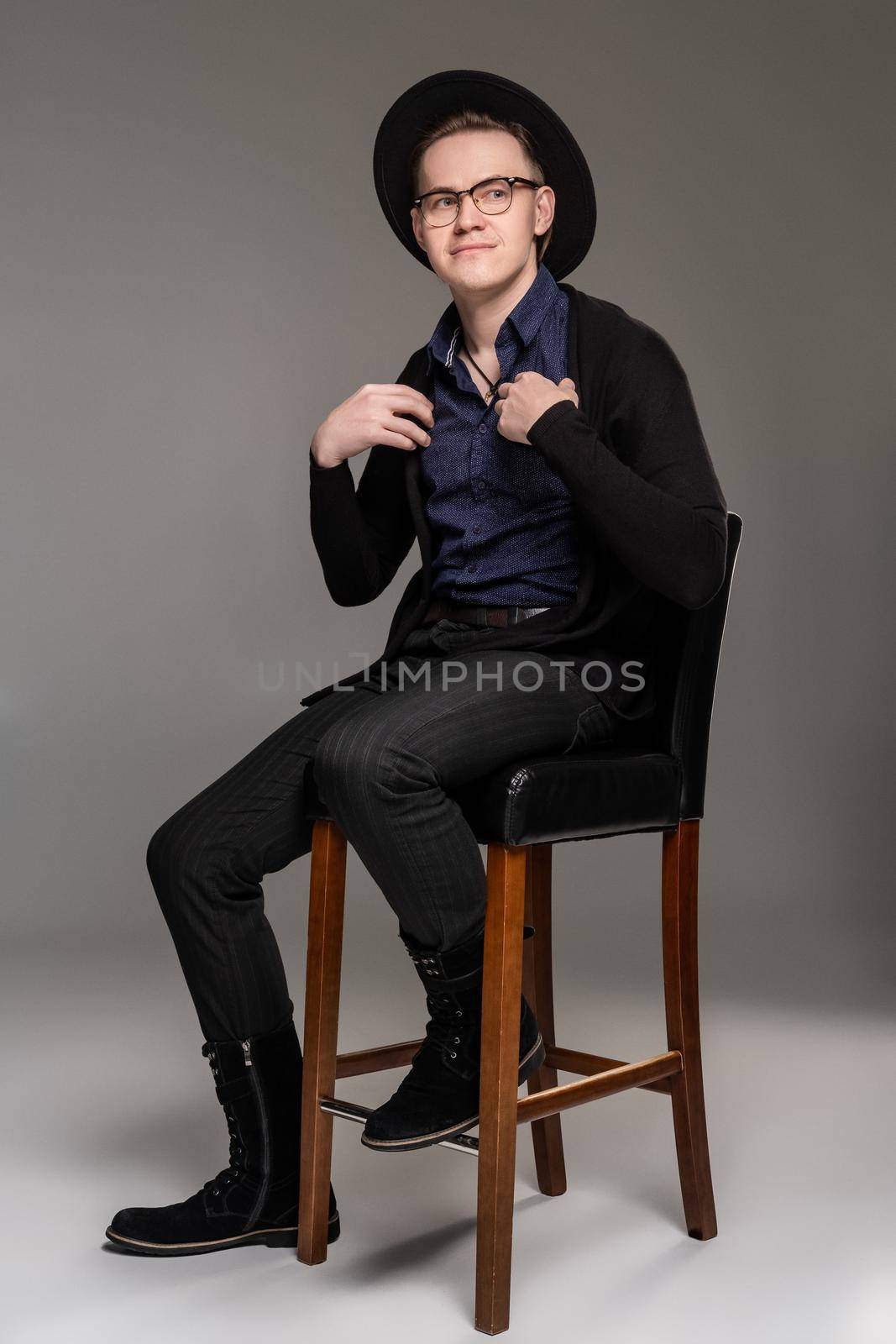Good-looking brunet guy in a casual dark clothes, stylish scarf, glases and black hat is sitting on the bar chair and looking at you while posing over a gray background. Copy space. Emotions concept.