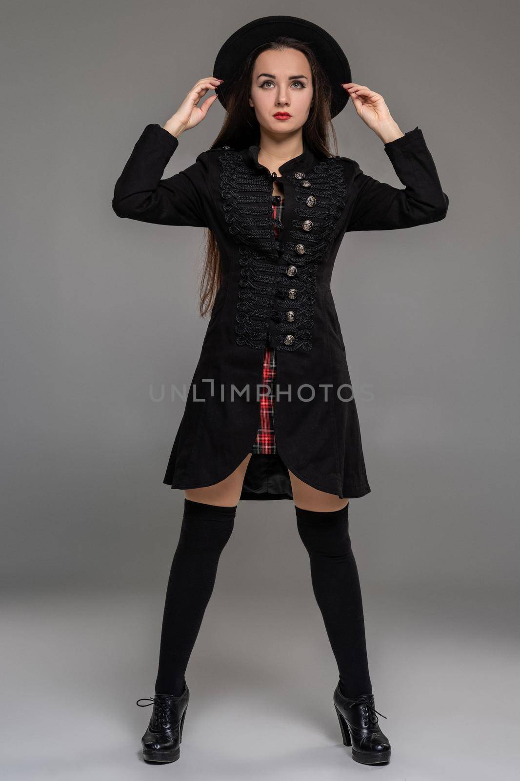 Full length portrait of an attractive female holding her hat and posing at studio against a gray background. She is weared in a checkered dress, black coat, stockings and boots. Fashion shot. Sincere emotions concept.