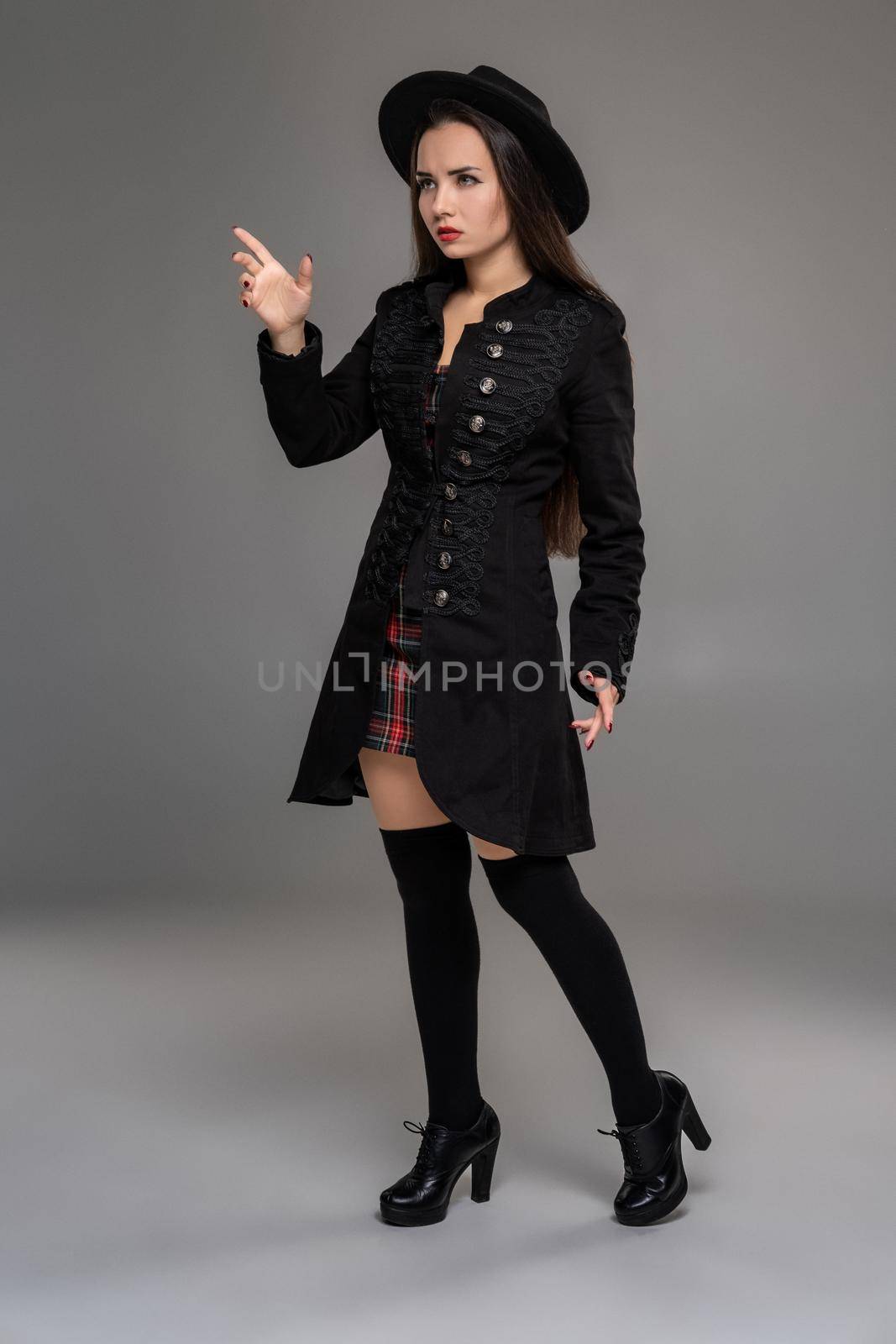 Full length portrait of a beautiful girl looking at someone and posing sideways at studio against a gray background. She is weared in a checkered dress, black coat, hat, stockings and boots. Fashion shot. Sincere emotions concept.