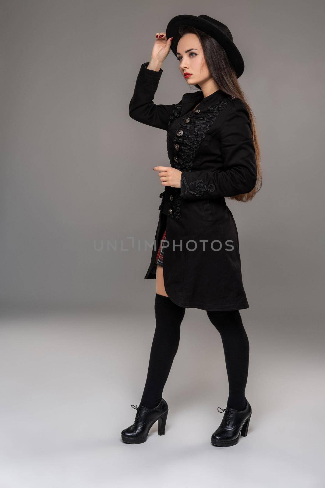 Full length portrait of a charming professional model posing sideways at studio against a gray background. She is weared in a checkered dress, black coat, hat, stockings and boots. Fashion shot. Sincere emotions concept.