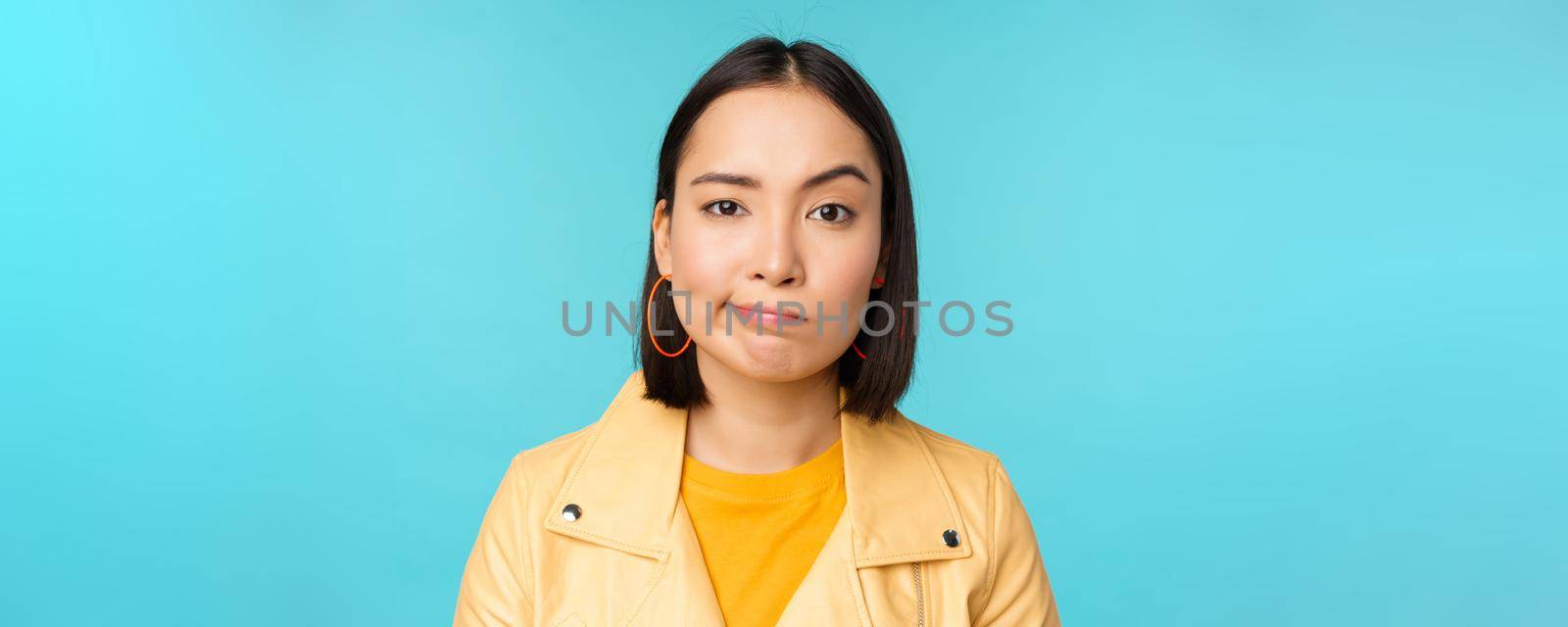 Close up of skeptical asian girl looking disappointed, staring with doubt or disbelief, grimacing at camera, standing over blue background.