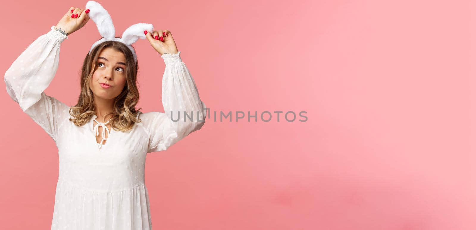 Holidays, spring and party concept. Portrait of silly cute blond girl in rabbit ears, looking up daydreaming, smiling lovely wear white dress, standing over pink background carefree by Benzoix