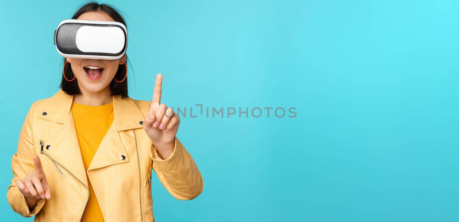 Young asian woman using virtual reality glasses, using VR headset, standing amused against blue background. Copy space