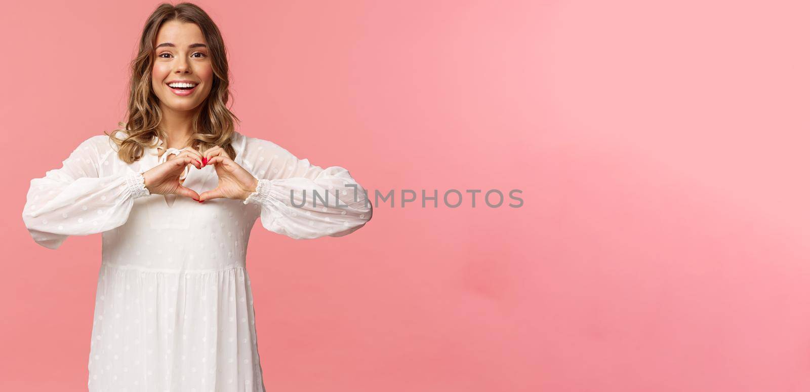 Beauty, fashion and women concept. Romantic passionate blond european female in white cute dress, make heart sign near chest and smiling at camera excited, show sympathy.