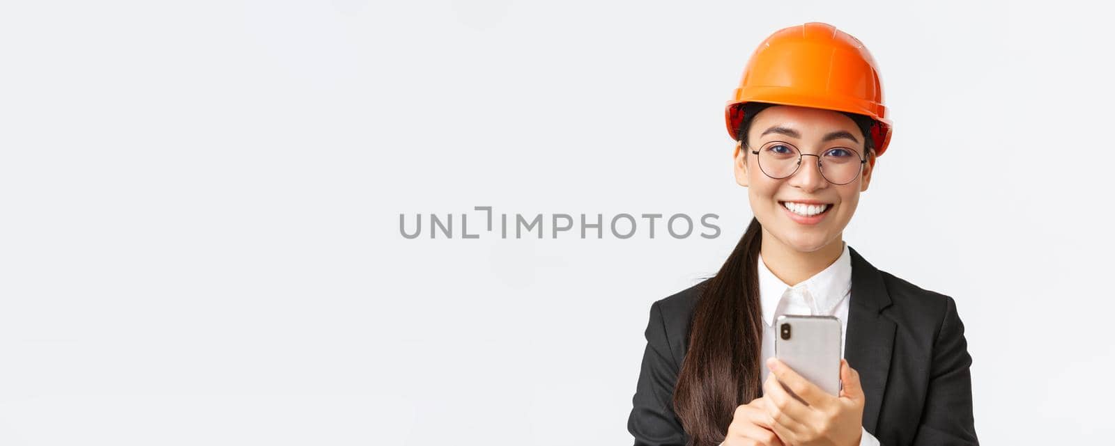 Close-up of successful female chief engineer, construction architect in safety helmet and business suit, glasses, looking at camera satisfied, smiling pleased as using mobile phone by Benzoix