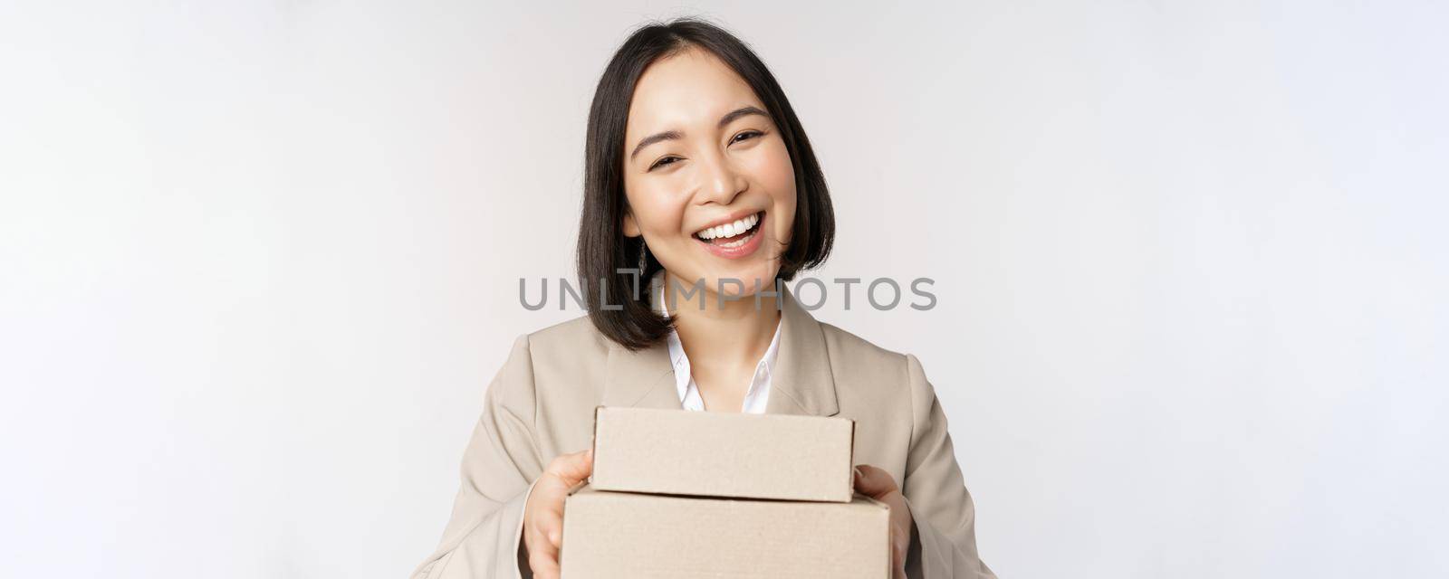 Image of asian saleswoman, business woman giving boxes with order, deliver to customer, standing in suit over white background.