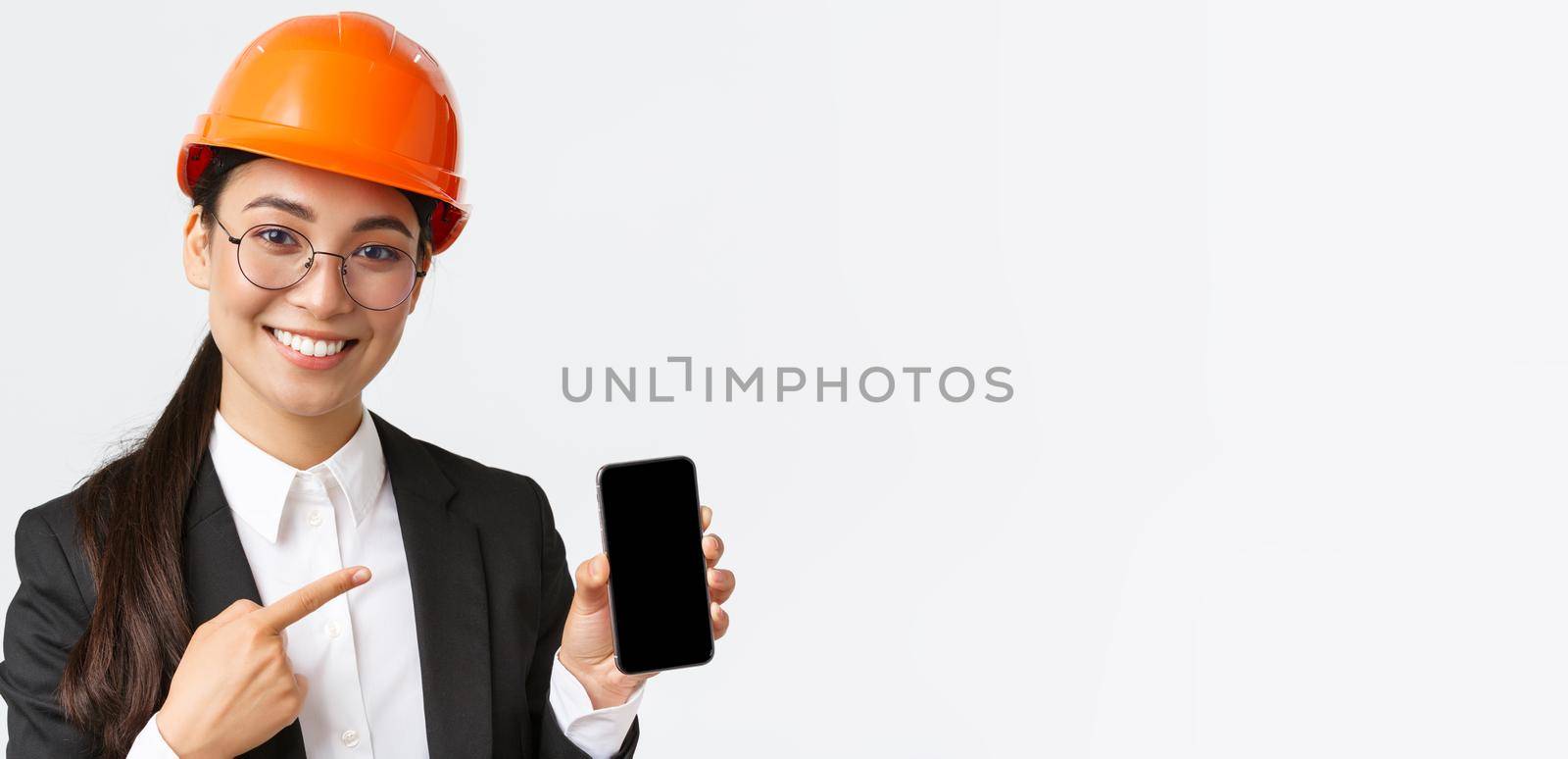 Close-up of smiling professional female asian construction engineer, architect in suit and safety mask introduce application, pointing finger at smartphone display, white background by Benzoix
