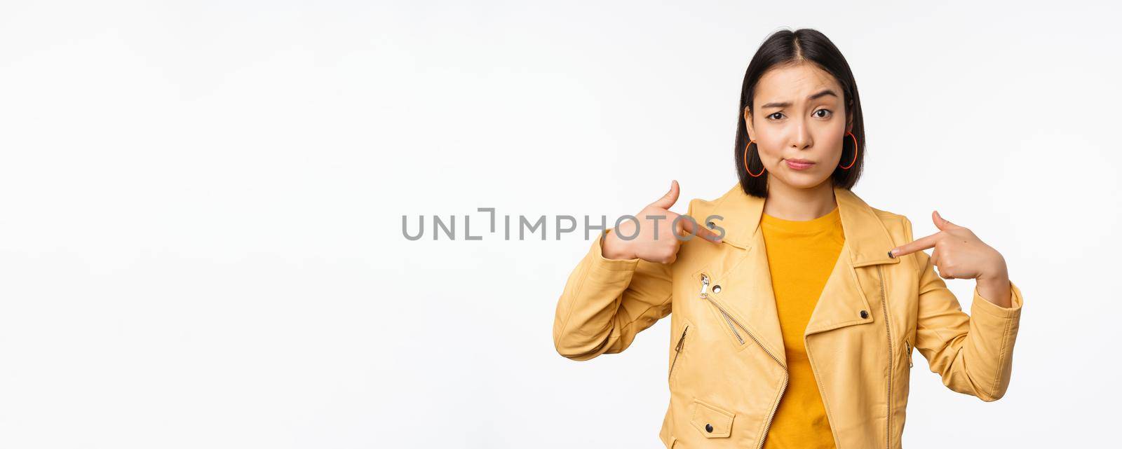 Image of unamused asian girl pointing at herself, looking with disbelief skeptical at camera, posing against white background.
