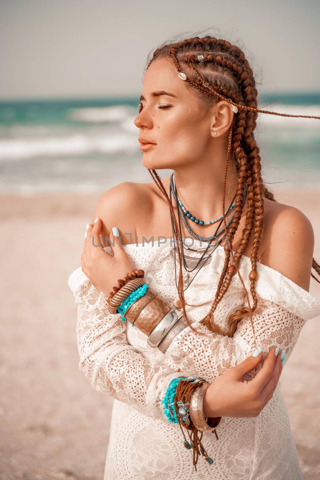 Model in boho style in a white long dress and silver jewelry on the beach. Her hair is braided, and there are many bracelets on her arms. by Matiunina