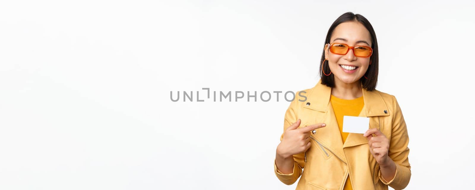 Portrait of beautiful modern asian girl in sunglasses, smiling happy, showing credit card, standing over white background by Benzoix