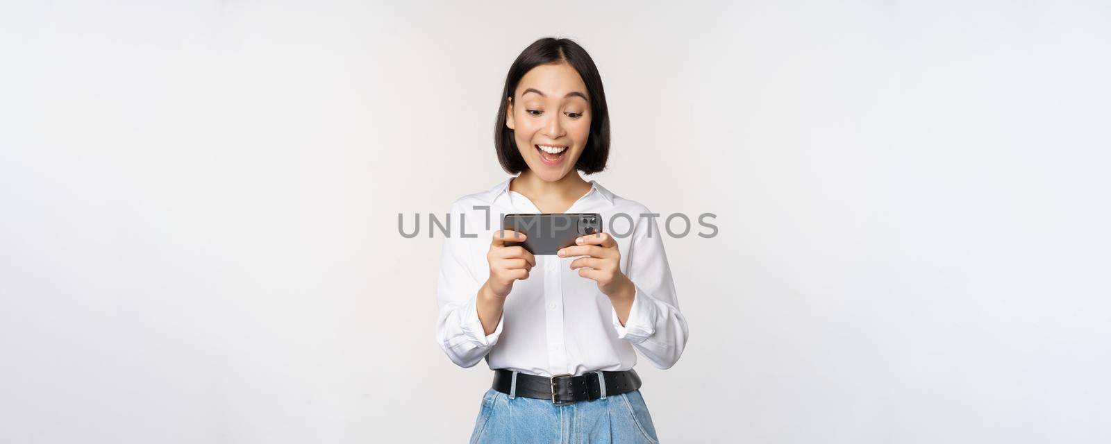 Image of young asian woman watching on smartphone app, holding mobile phone and looking at screen, laughing and smiling, standing over white background.