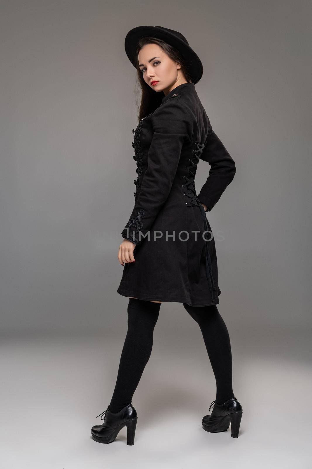 Full length portrait of an attractive girl looking at the camera and posing back at studio against a gray background. She is weared in a checkered dress, black coat, hat, stockings and boots. Fashion shot. Sincere emotions concept.