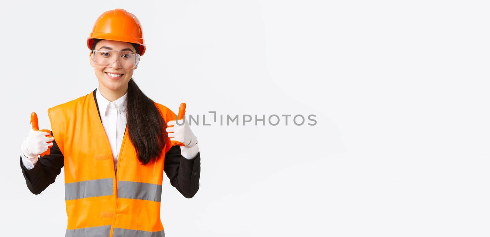Satisfied smiling female asian chief construction engineer giving permission to enter building or entertprise after wearing safety clothing, glasses gloves and helmet, showing thumbs-up in approval.