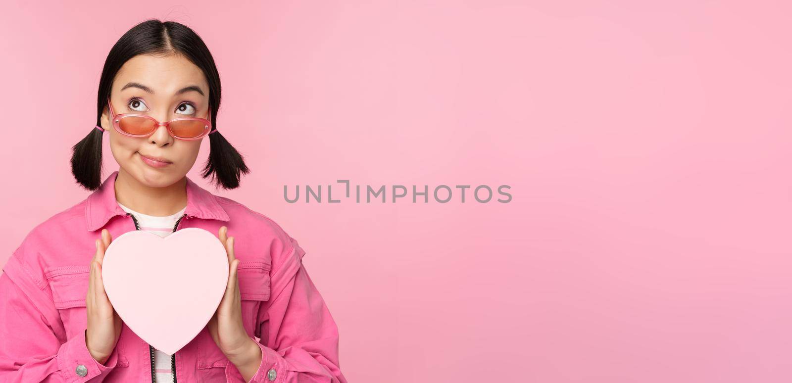 Beautiful asian girl smiling happy, showing heart gift box and looking excited at camera, standing over pink romantic background by Benzoix