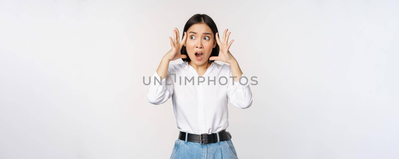 Image of shocked anxious asian woman in panic, holding hands on head and worrying, standing frustrated and scared against white background.