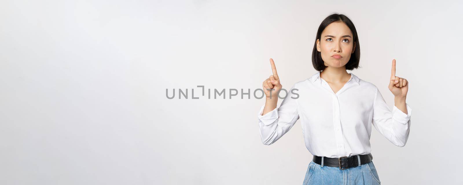 Skeptical businesswoman, asian office manager, pointing fingers up and grimacing doubtful, hesitating, standing over white background.