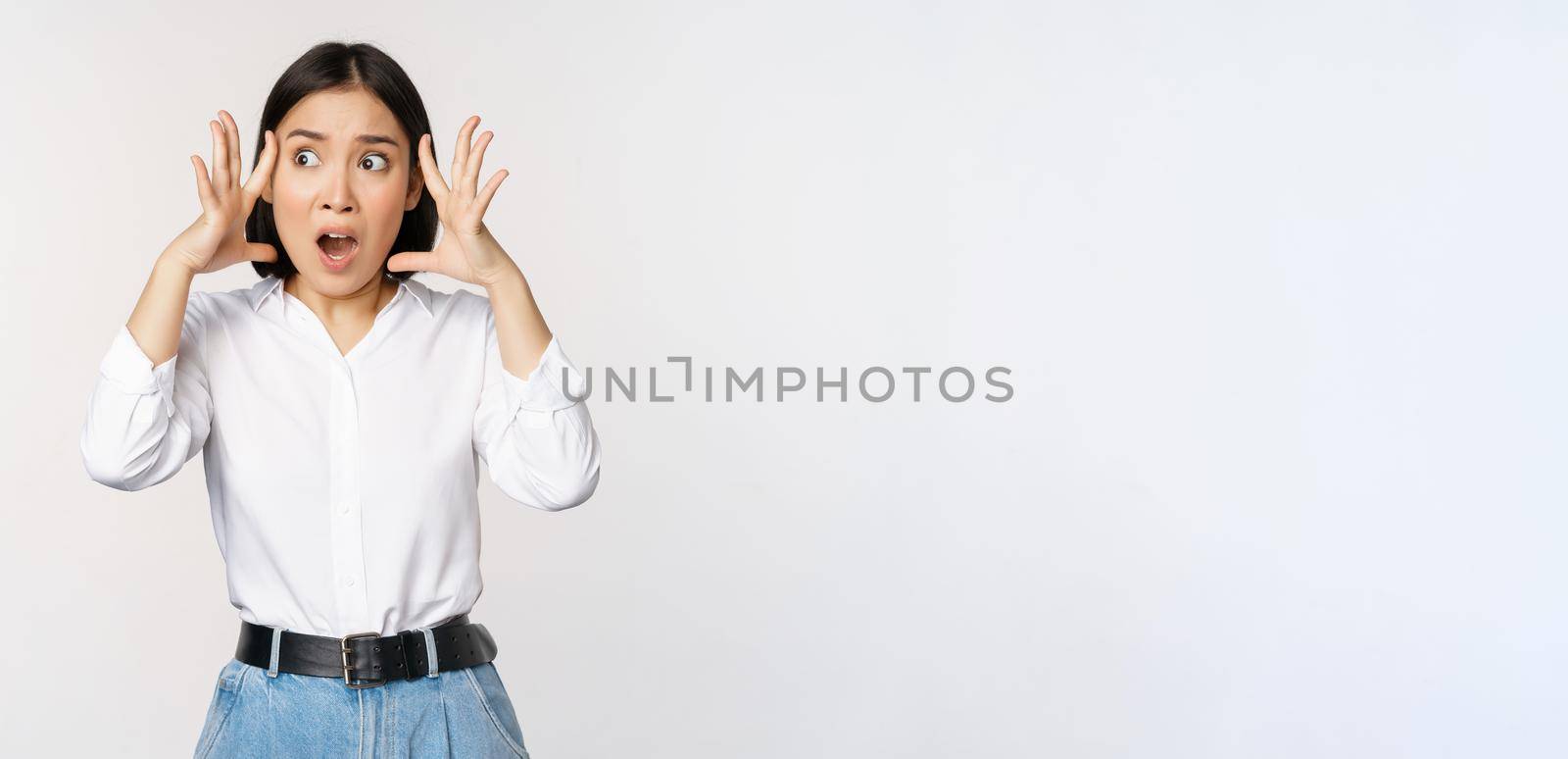 Image of shocked anxious asian woman in panic, holding hands on head and worrying, standing frustrated and scared against white background.