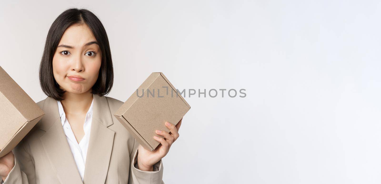 Puzzled asian saleswoman holding boxes, looking confused, standing in business suit over white background. Copy space