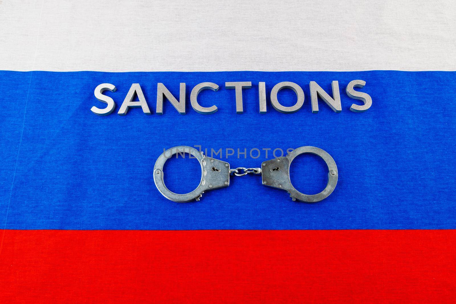 the word sanctions laid with silver metal letters on russian tricolor flag near handcuffs over it in directly above view